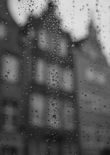 Black and white photo of raindrops on a window with blurred urban background, creating a moody atmosphere.