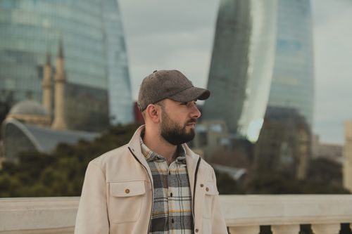 Free A man with a beard and cap stands in front of modern glass towers in an urban environment. Stock Photo