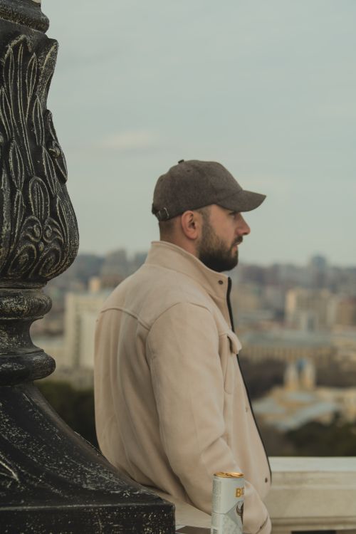 Free A man in jacket and cap overlooks a cityscape while holding a beer can. Stock Photo