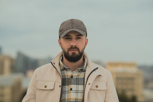 Free Portrait of a man in a cap and plaid shirt against an outdoor cityscape backdrop. Stock Photo