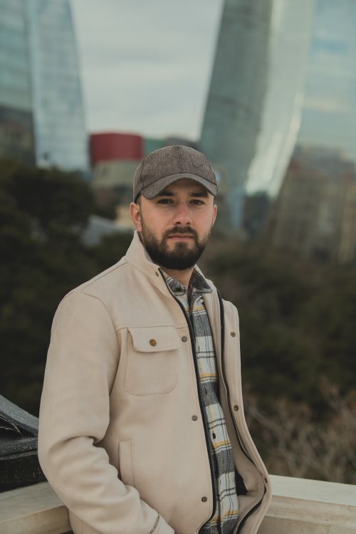 Free Bearded man in a cap and jacket posing outdoors with cityscape in the background. Stock Photo
