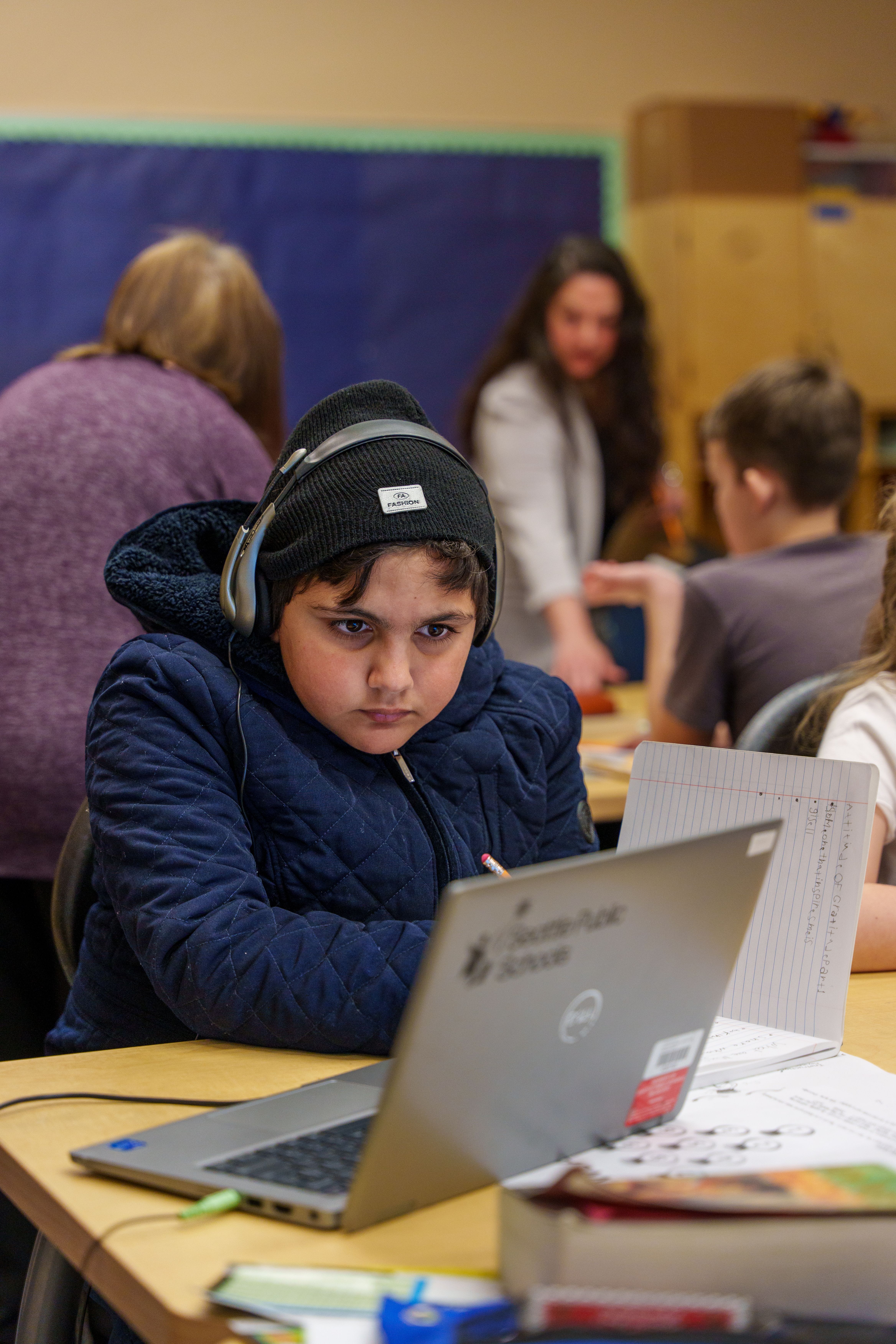 Young student wearing headphones focuses intently on a laptop in a busy classroom setting.