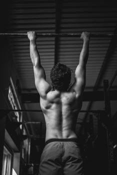Black and white image of a shirtless man doing a pull-up in a gym, showcasing strength and fitness.