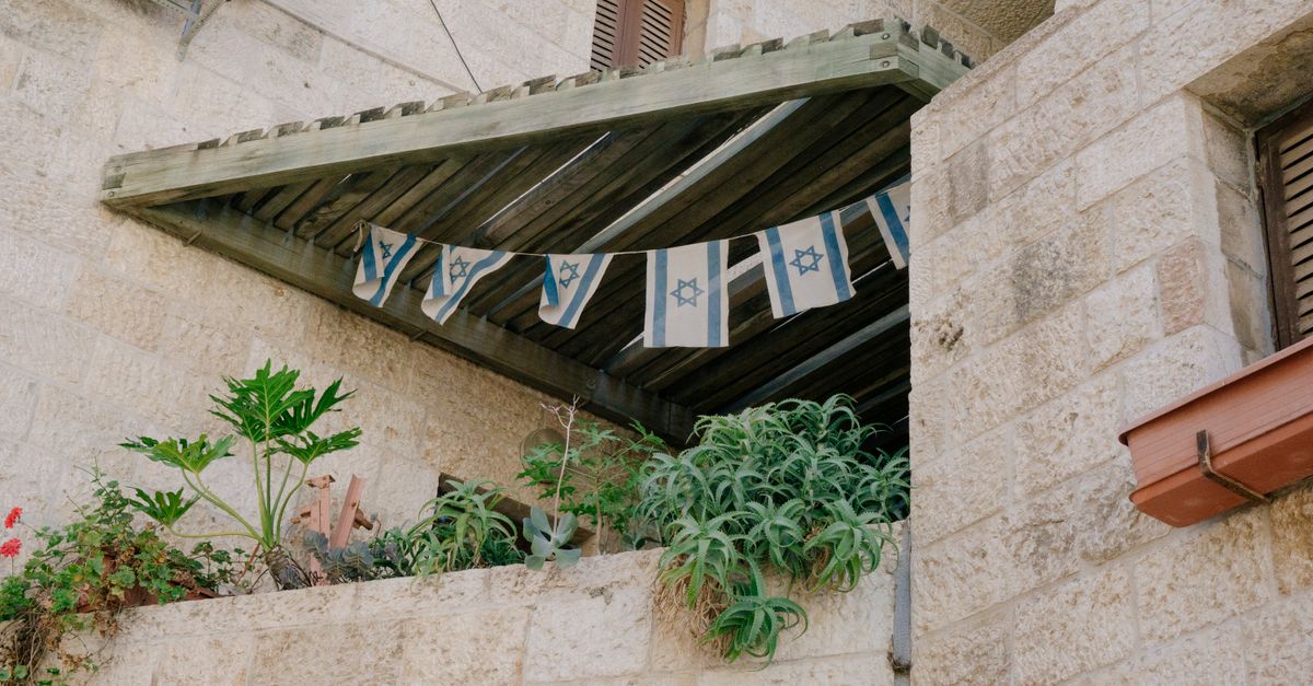 Stone building exterior with Israeli flags and diverse plants on balcony in urban setting.