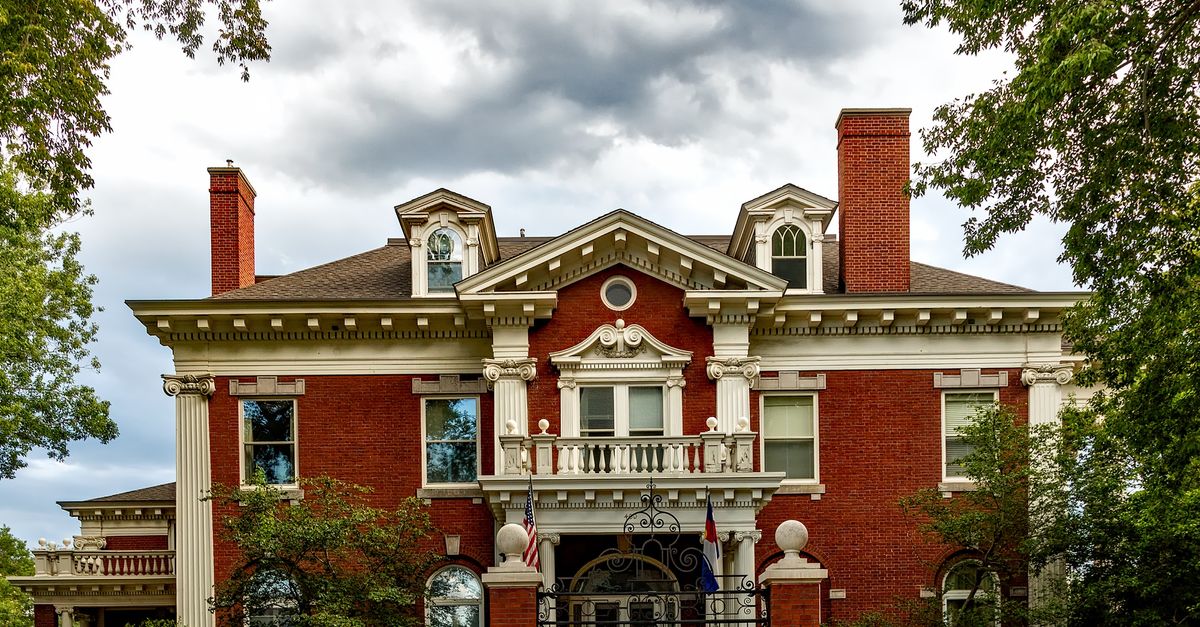 Historic red brick mansion with a grand entrance and ornate gate under a cloudy sky.