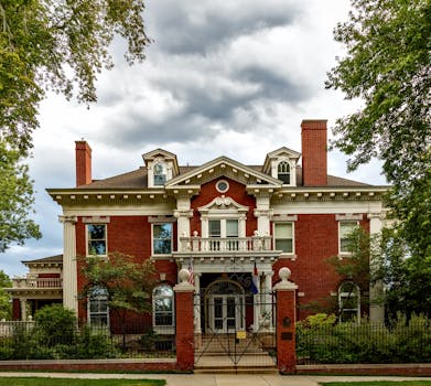 Historic red brick mansion with a grand entrance and ornate gate under a cloudy sky.