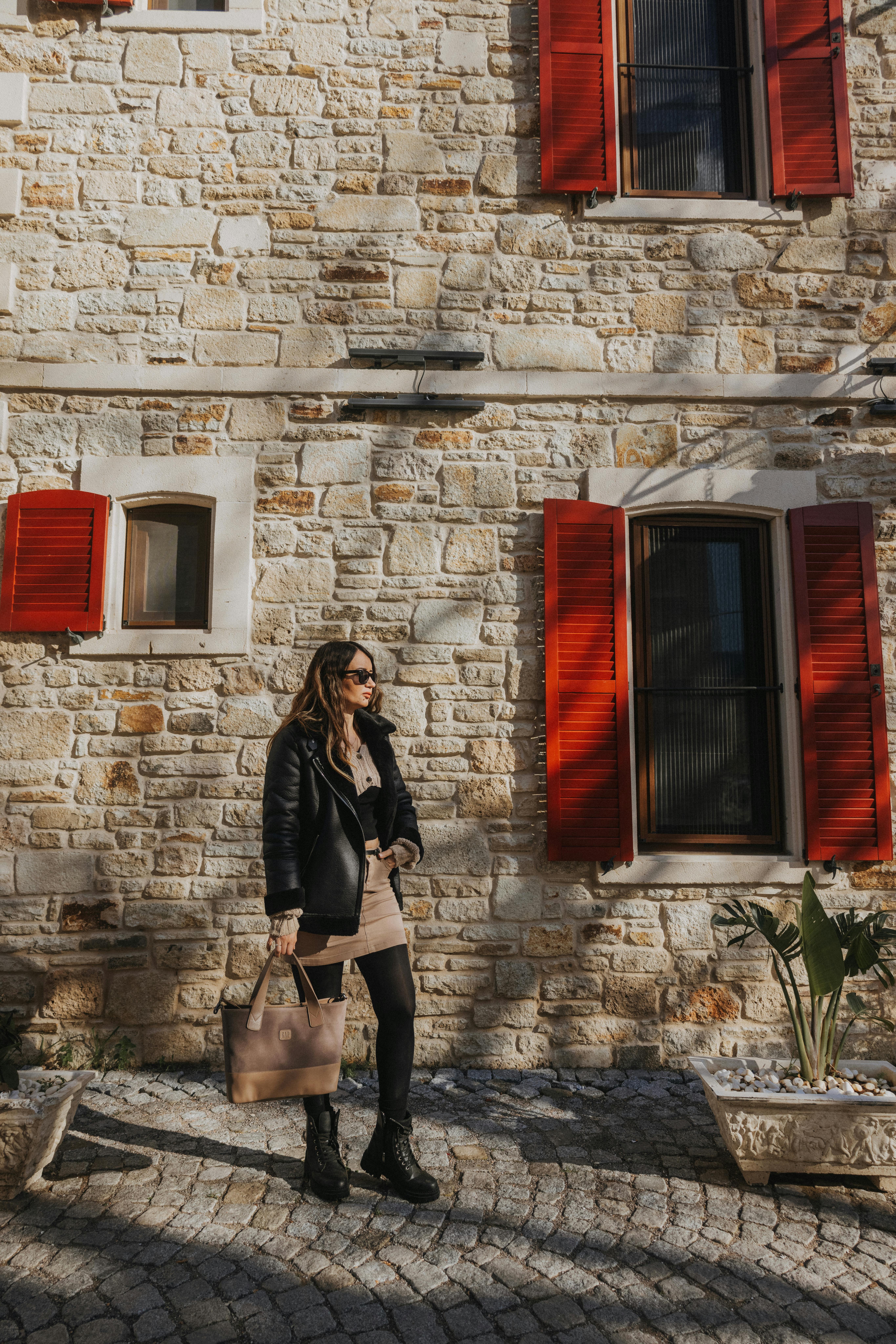 A stylish woman stands against a stone building with vibrant red window shutters, enjoying a sunny urban day.