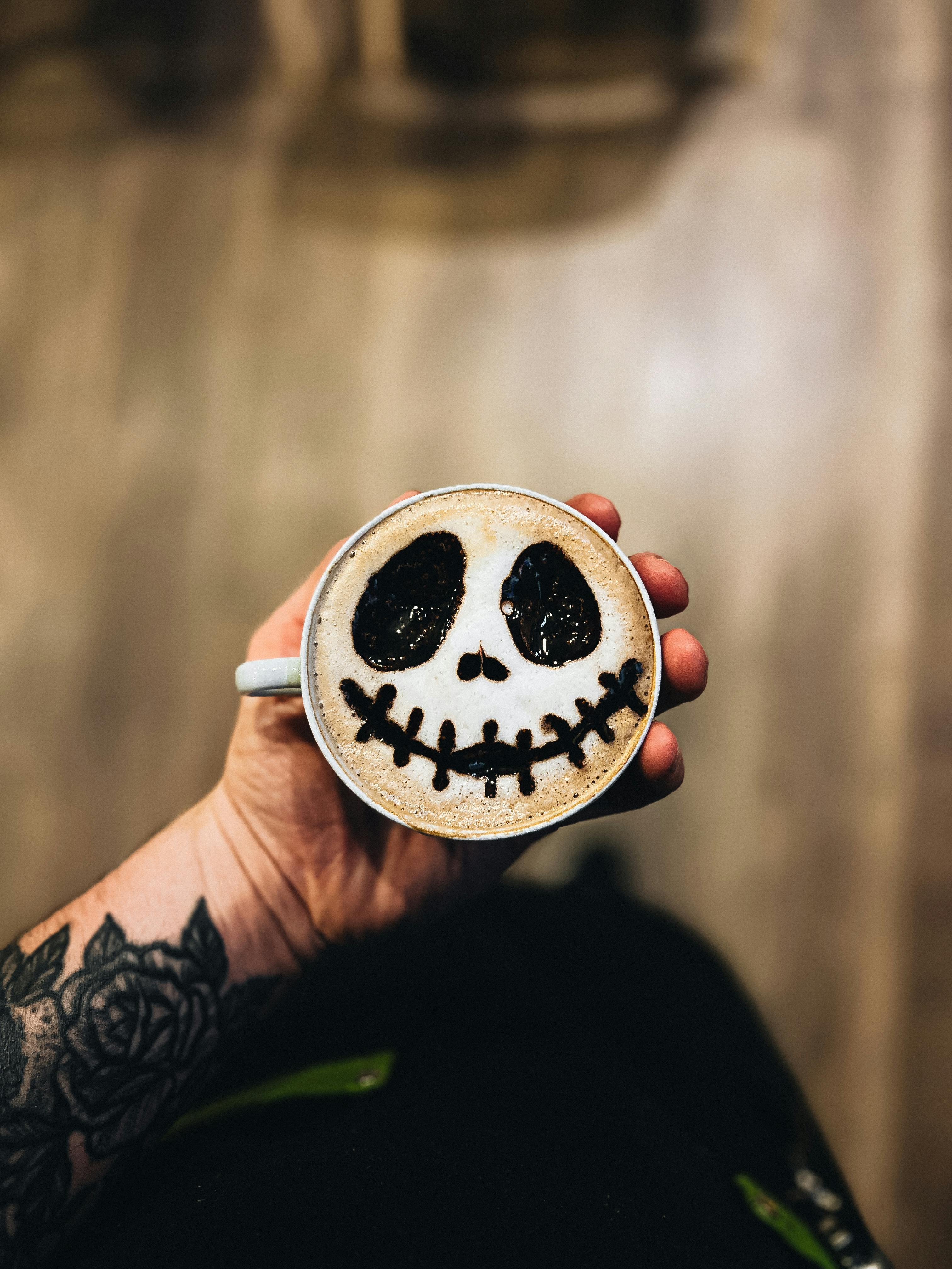 Creative latte art with a spooky Halloween face design in a cup held by a tattooed hand.