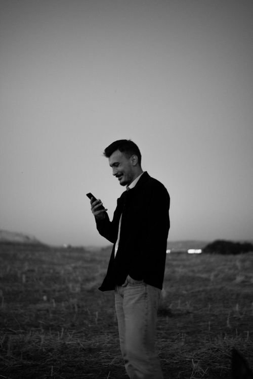 Free A man in a blazer checks his smartphone in an open field during sunset. Stock Photo