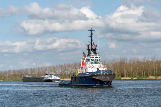 A tugboat and a cargo ship sailing on a calm river with a tree-lined background under a partly cloudy sky.