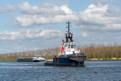 Free A tugboat and a cargo ship sailing on a calm river with a tree-lined background under a partly cloudy sky. Stock Photo