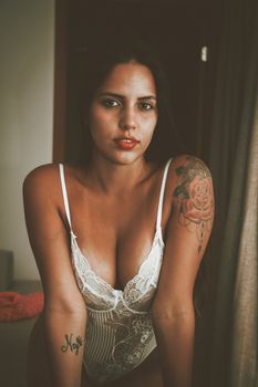 Fashion portrait of a woman in white lingerie showcasing tattoos in a serene indoor setting.