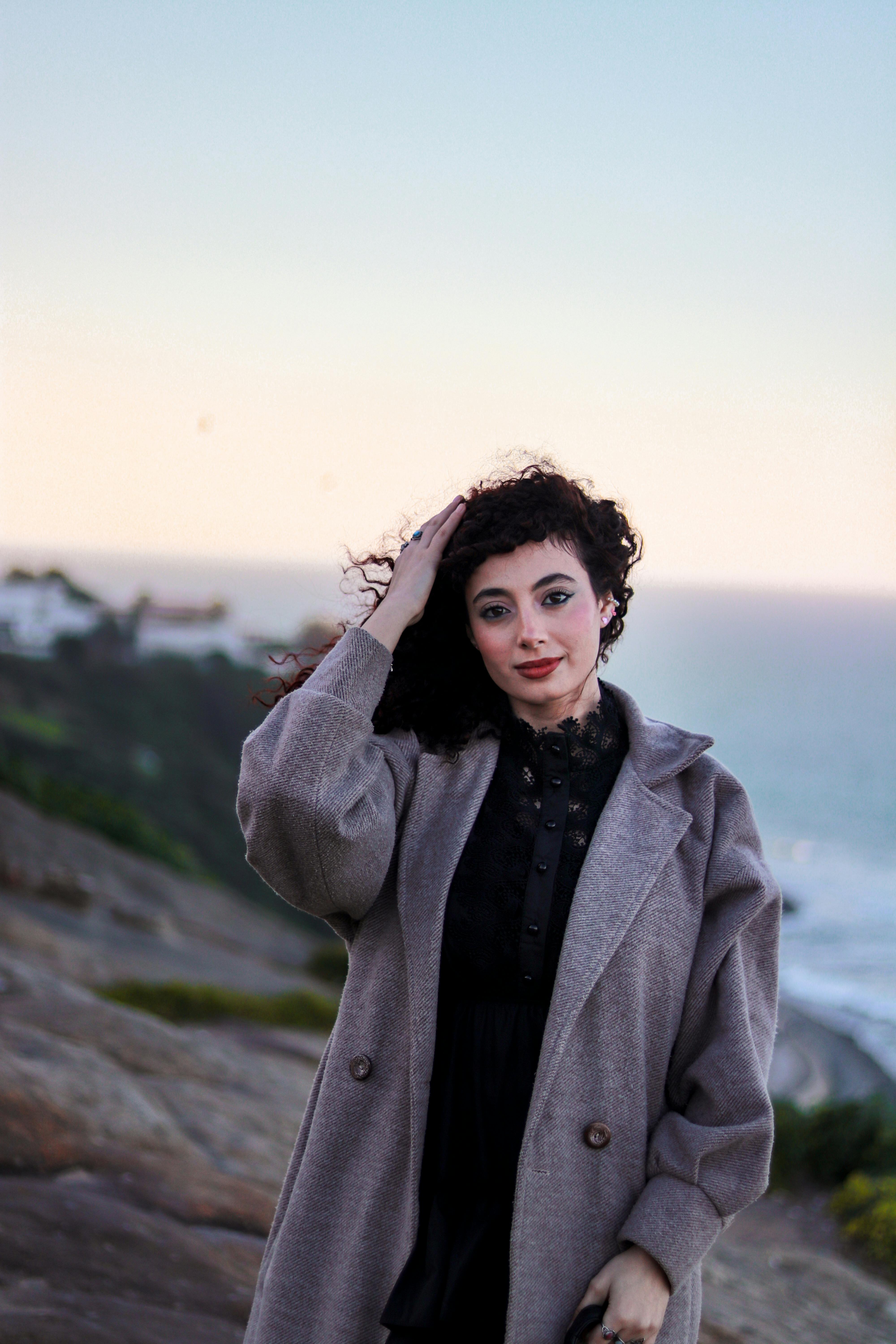 A stylish woman in a gray coat poses by the ocean at sunset, exuding elegance and confidence.