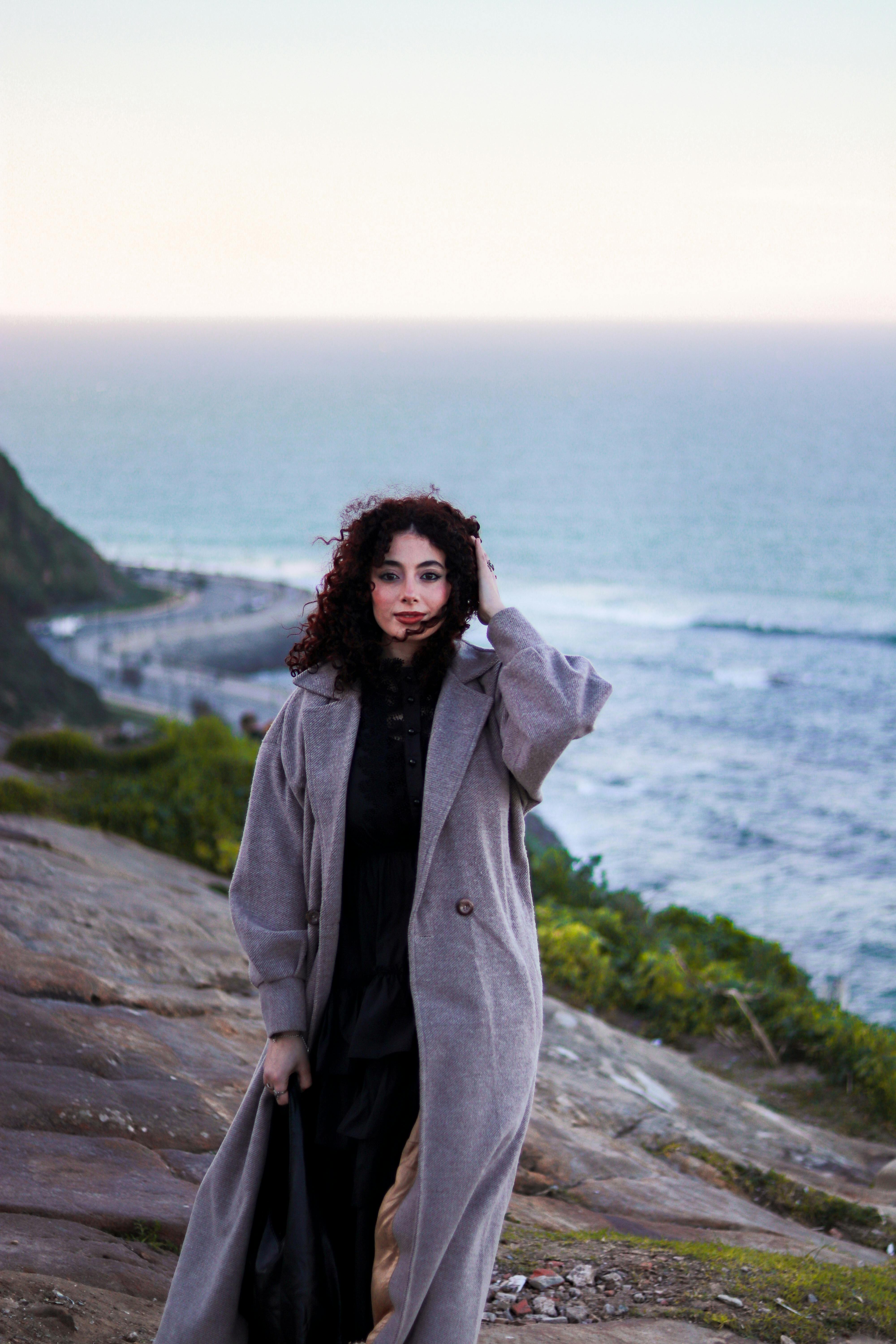 Woman with curly hair in a coat stands by the ocean at twilight, capturing the serene coastal scenery.