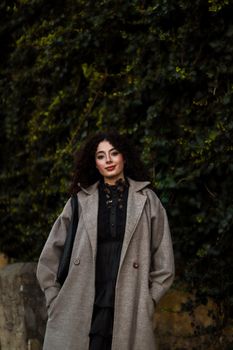 Glamorous woman in fashionable attire posing against lush greenery background.