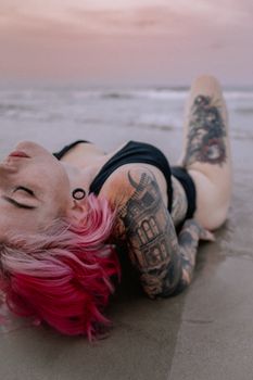 A woman with vibrant tattoos and pink hair, posing on a sandy beach during sunset.
