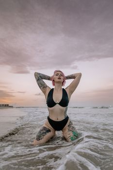 A tattooed woman poses in a bikini on the beach at sunset, surrounded by waves.