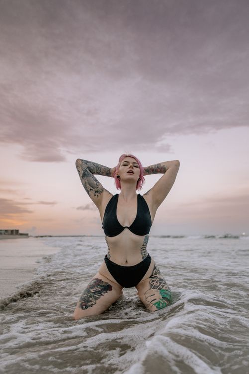 Free A tattooed woman poses in a bikini on the beach at sunset, surrounded by waves. Stock Photo