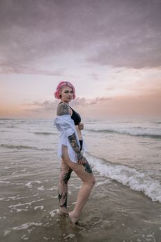 A vibrant woman with tattoos and pink hair stands on a beach at sunset, exuding confidence and style.