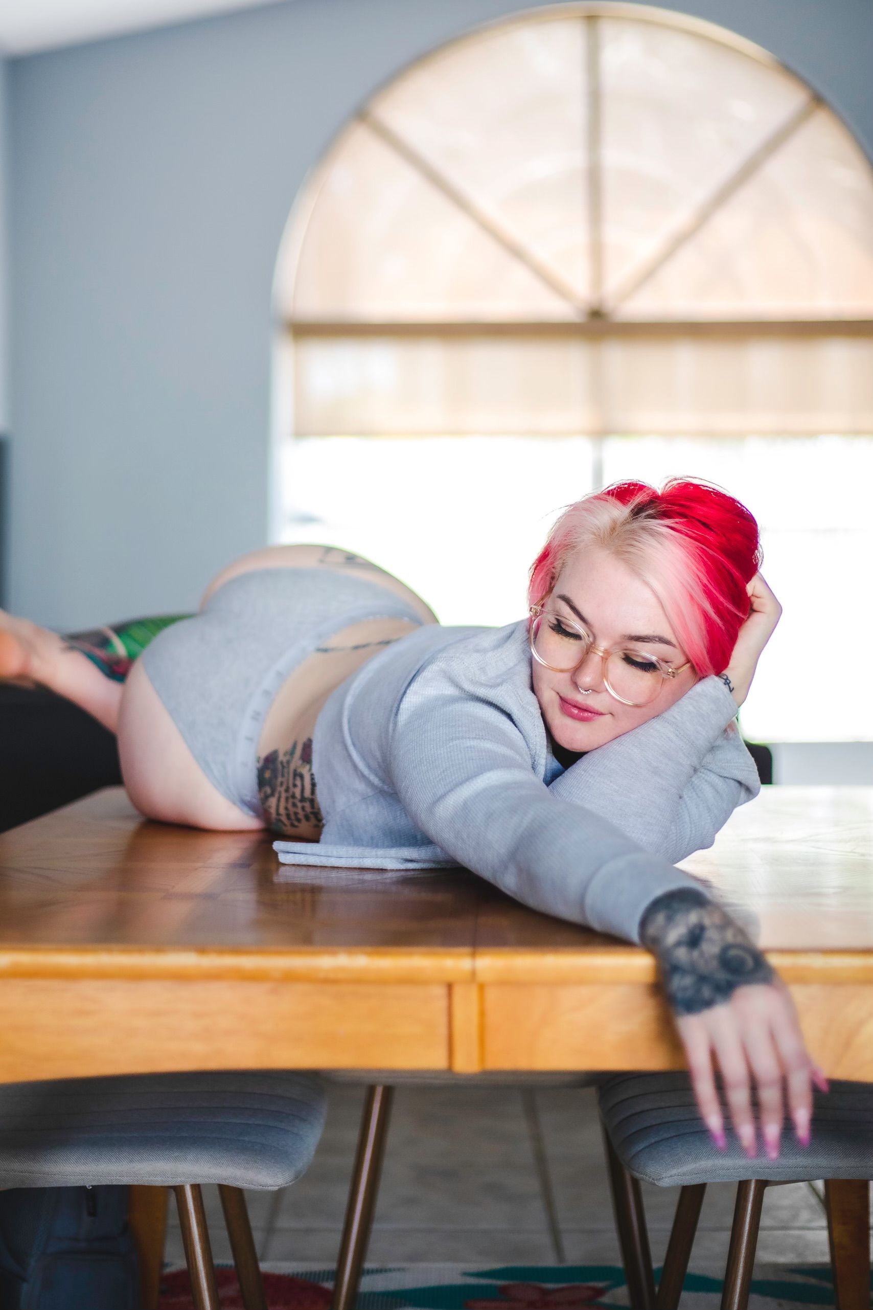Fashion photography of a young woman with tattoos and pink hair posing on a table indoors.