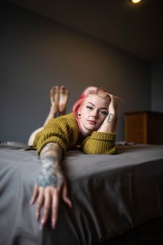 Portrait of a tattooed woman with pink hair lying on a bed in a cozy, dimly lit bedroom.