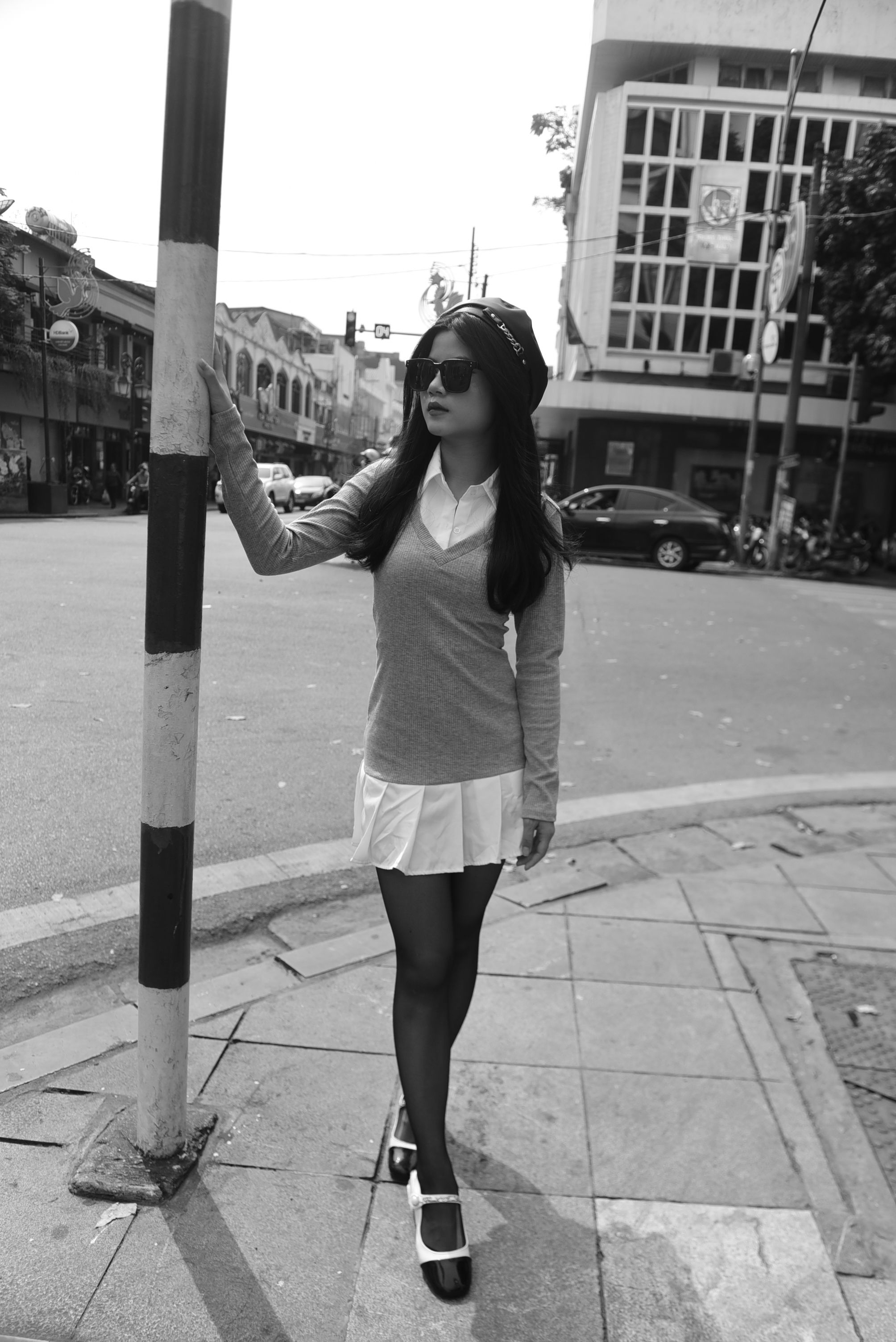 A fashionable woman leans on a pole on a city sidewalk, striking an urban pose in black and white.