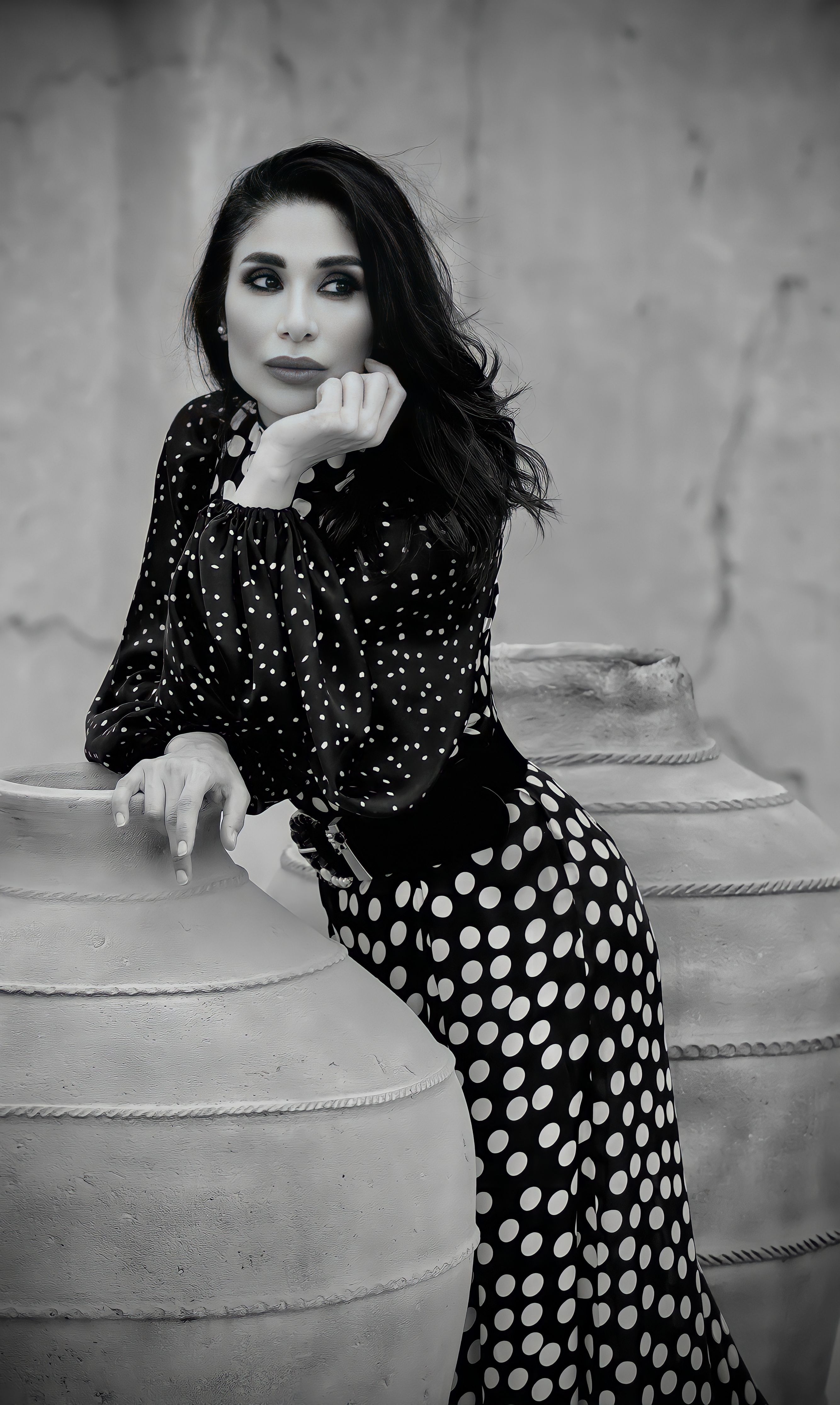 Black and white portrait of a woman in polka dot attire leaning against rustic vases.
