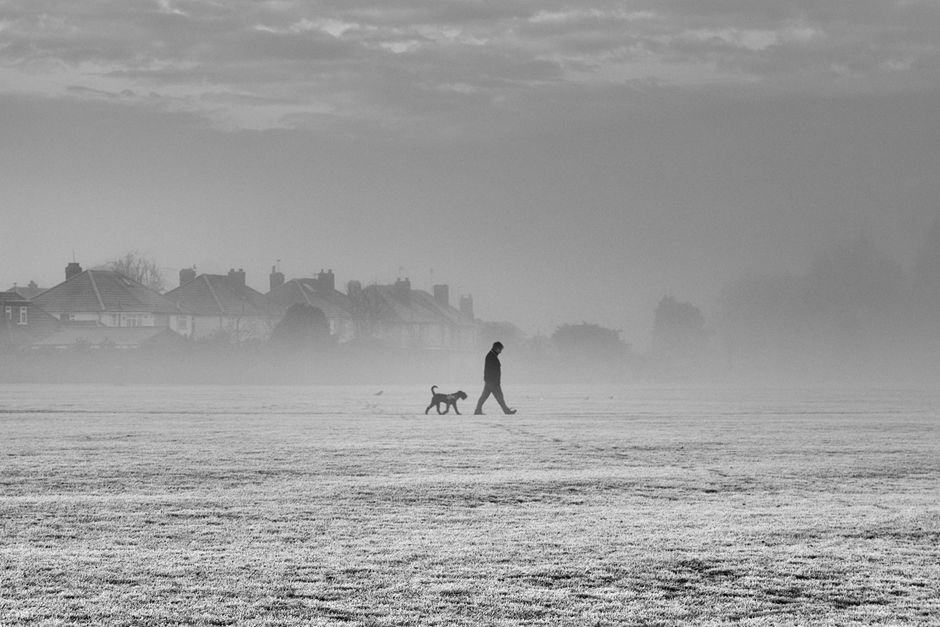 A silhouette of a man walking his dog through a foggy park in England.