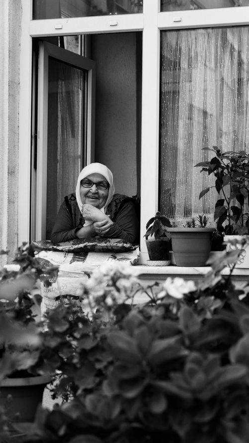 Free Black and white photo of a smiling elderly woman leaning out a window surrounded by plants. Stock Photo