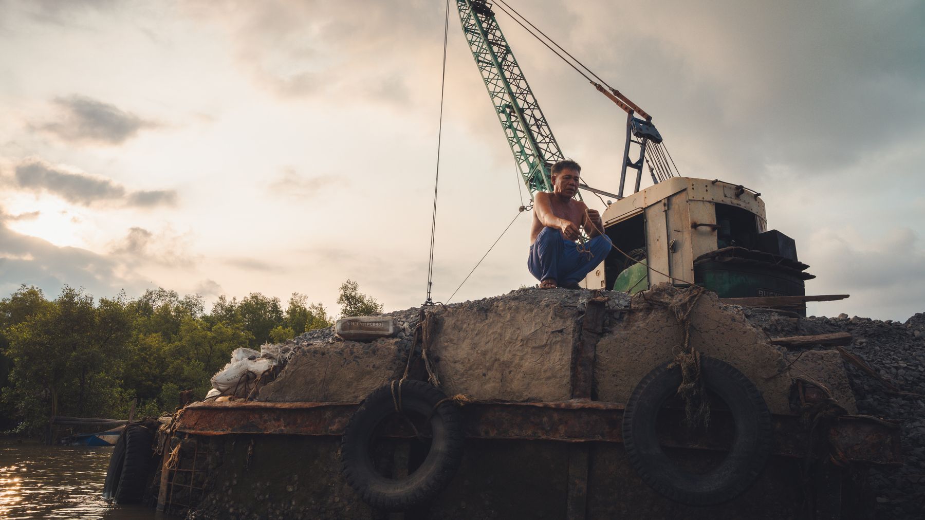 A man sits on a construction barge in a canal in Gò Công, Vietnam, at sunset.