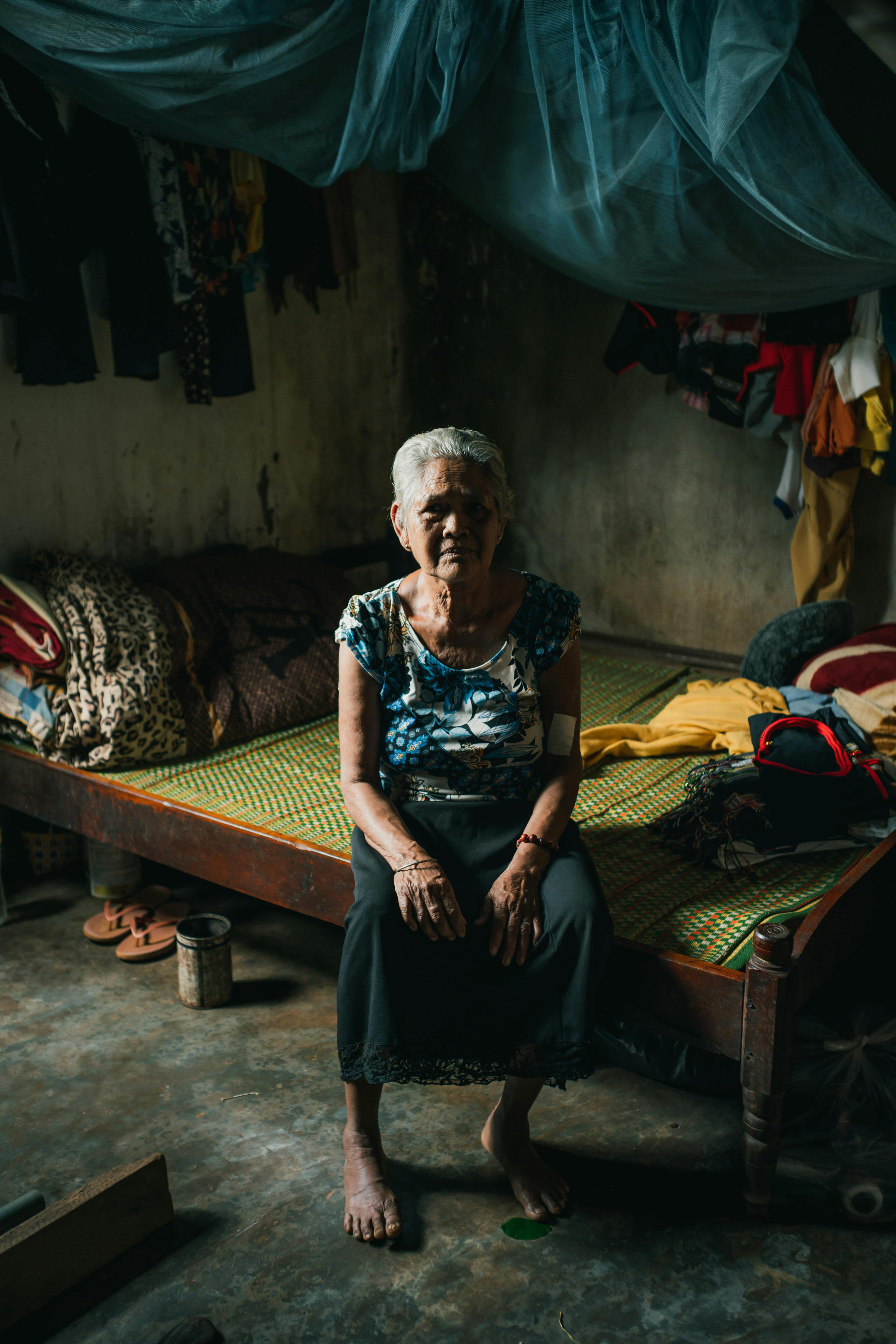 Elderly woman seated on a bed in a rustic, dimly-lit room in Vietnam.