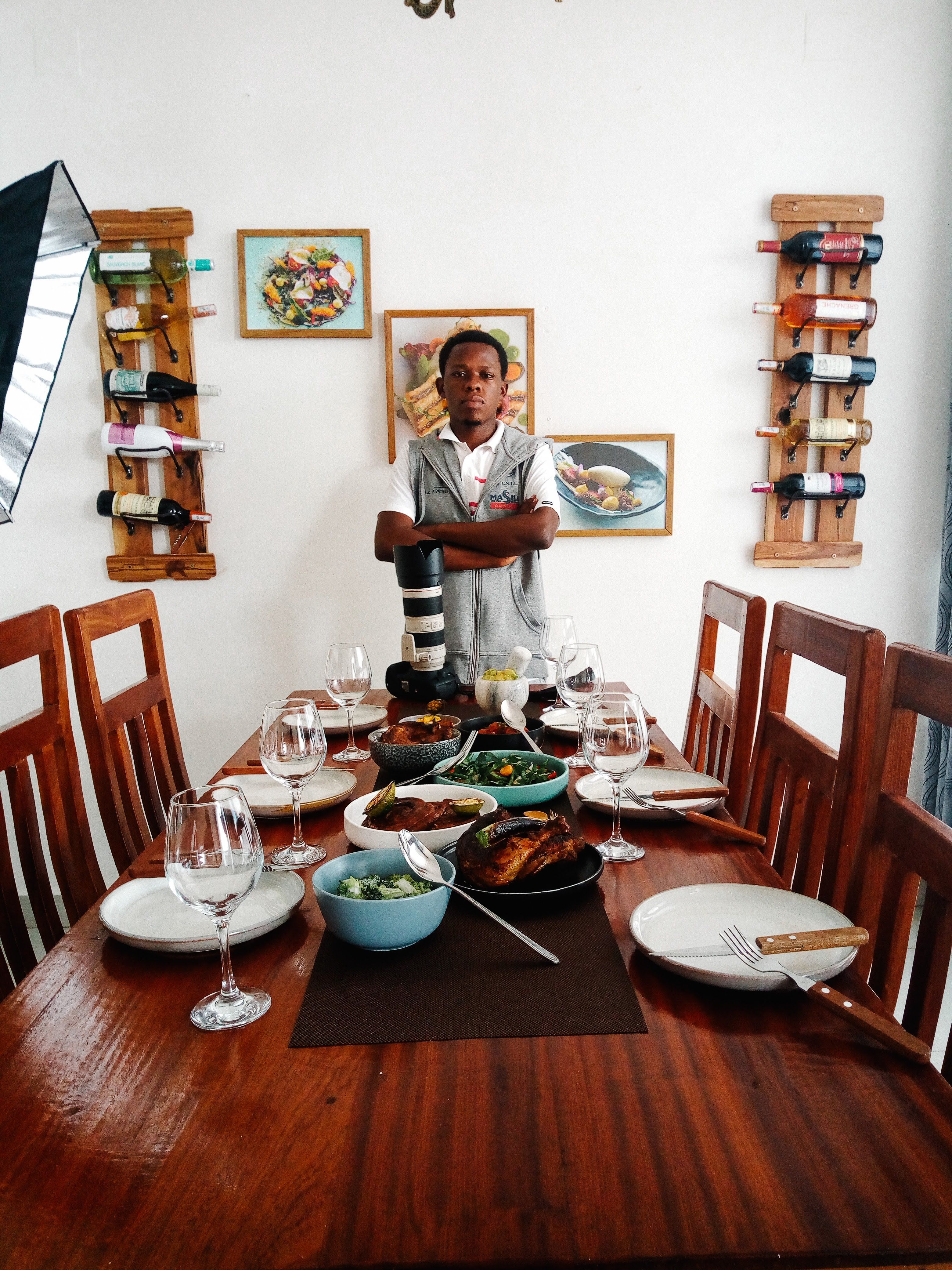 Table de restaurant dressée : verres à pied, assiettes, bols de légumes verts et de ragoût, poisson grillé au centre, un homme en tablier debout au fond