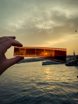 A hand holds a film negative against a stunning sunset by the water, capturing the serene scene.