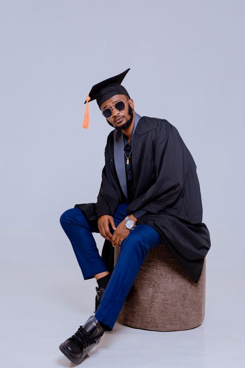 A man in graduation cap and gown sitting on a stool