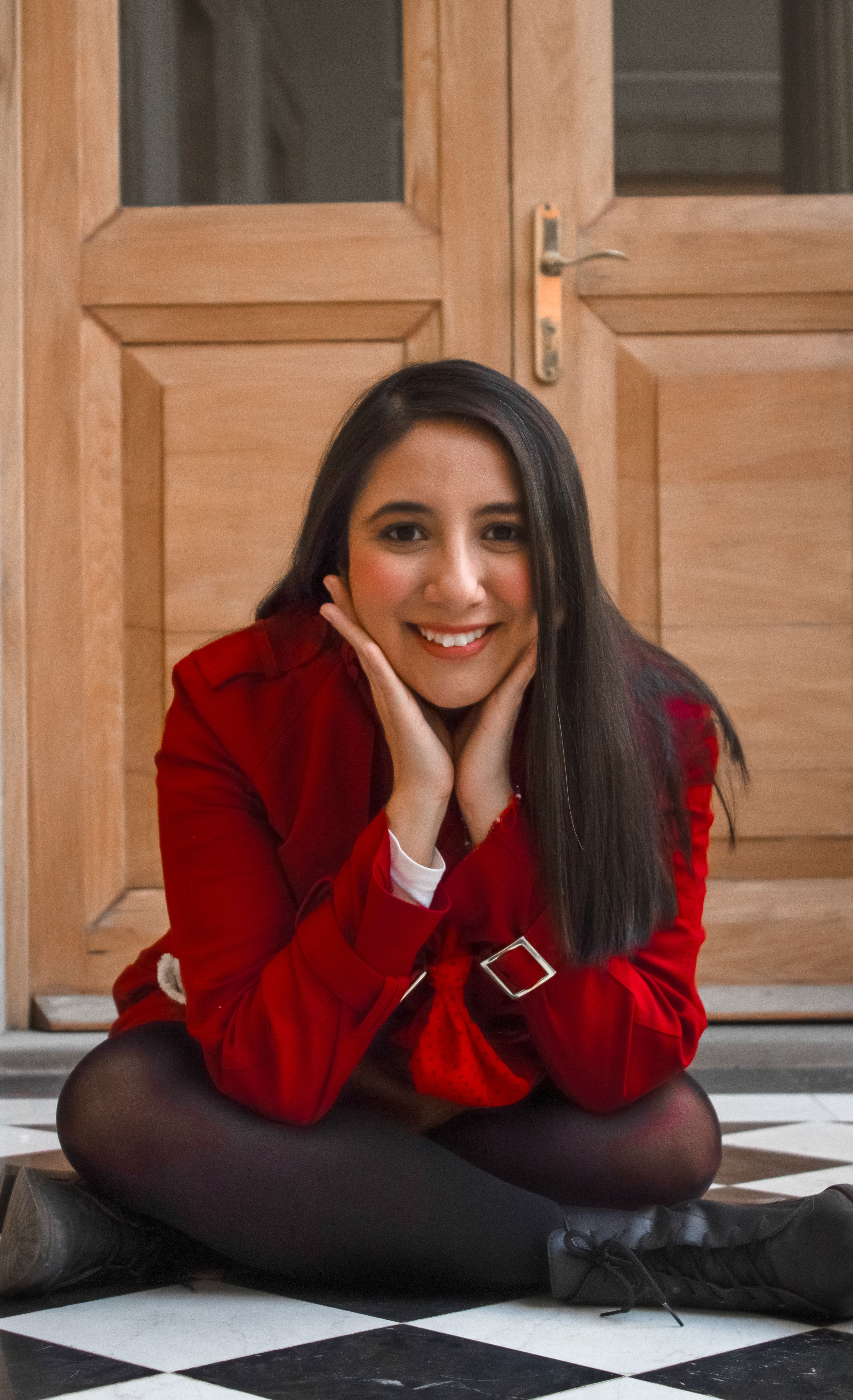 Portrait of woman sitting on floor with hands on cheeks, wearing red jacket and smiling.