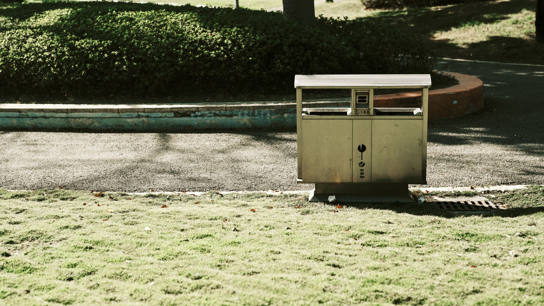 A solitary garbage bin in a quiet park surrounded by greenery and pavement.