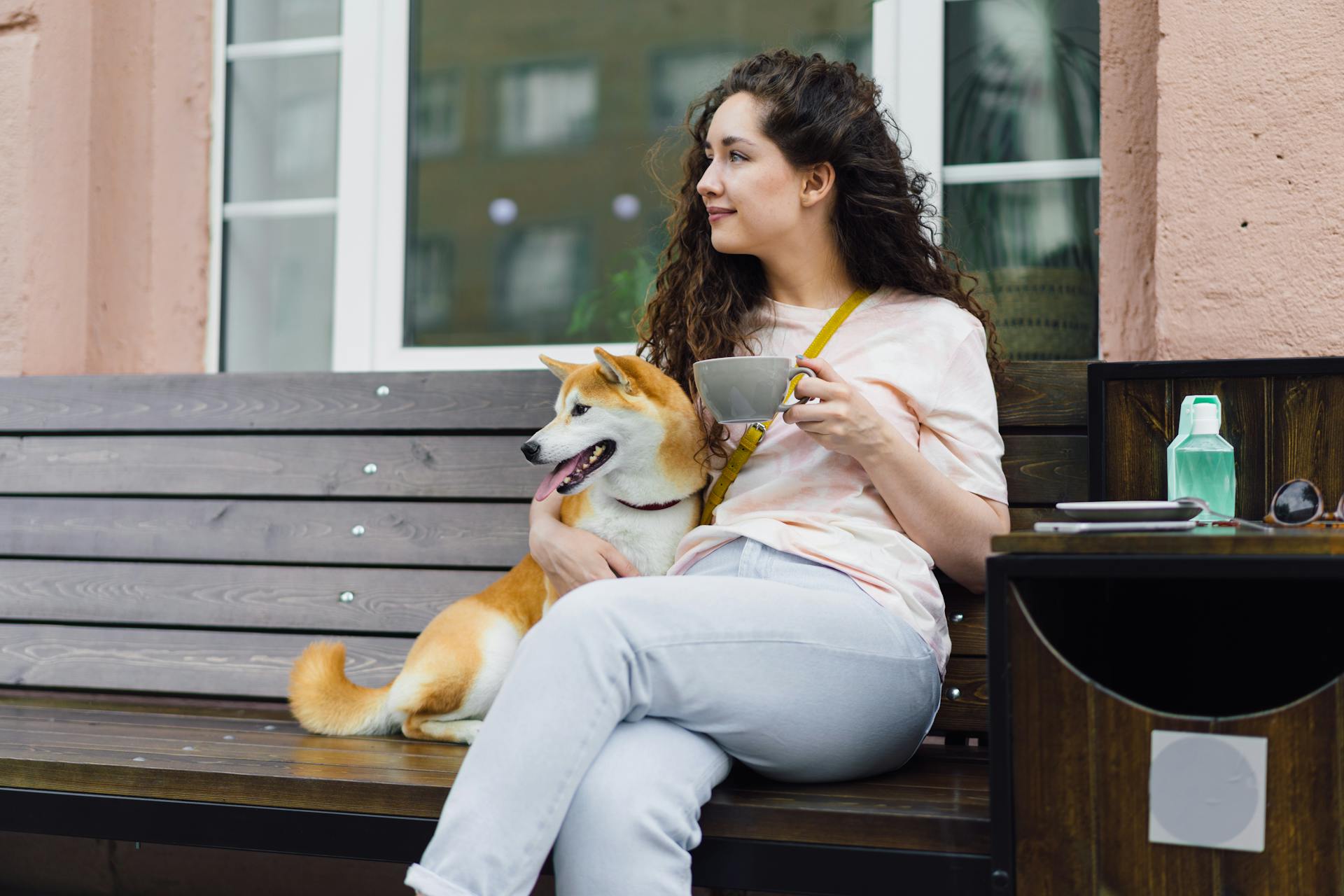 En kvinna sitter på en bänk med en kopp kaffe och en shiba-inu-hund