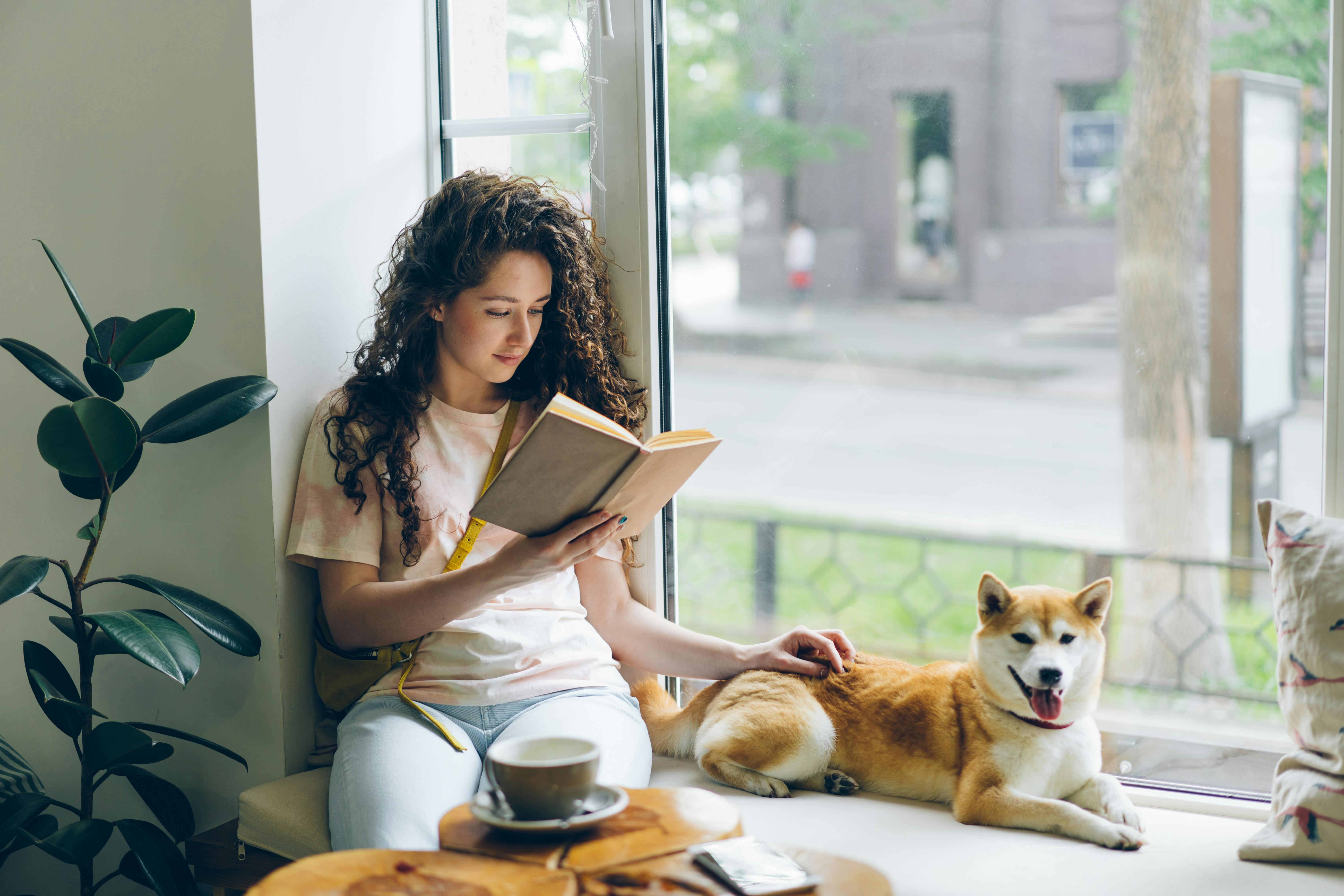 A Woman Reading a Book while Sitting on a Windowsill with Her Shiba Inu Dog in a Cafe