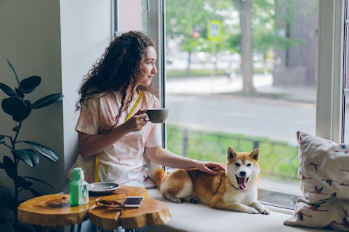 A woman holding a cup of coffee and a dog sitting on the window sill