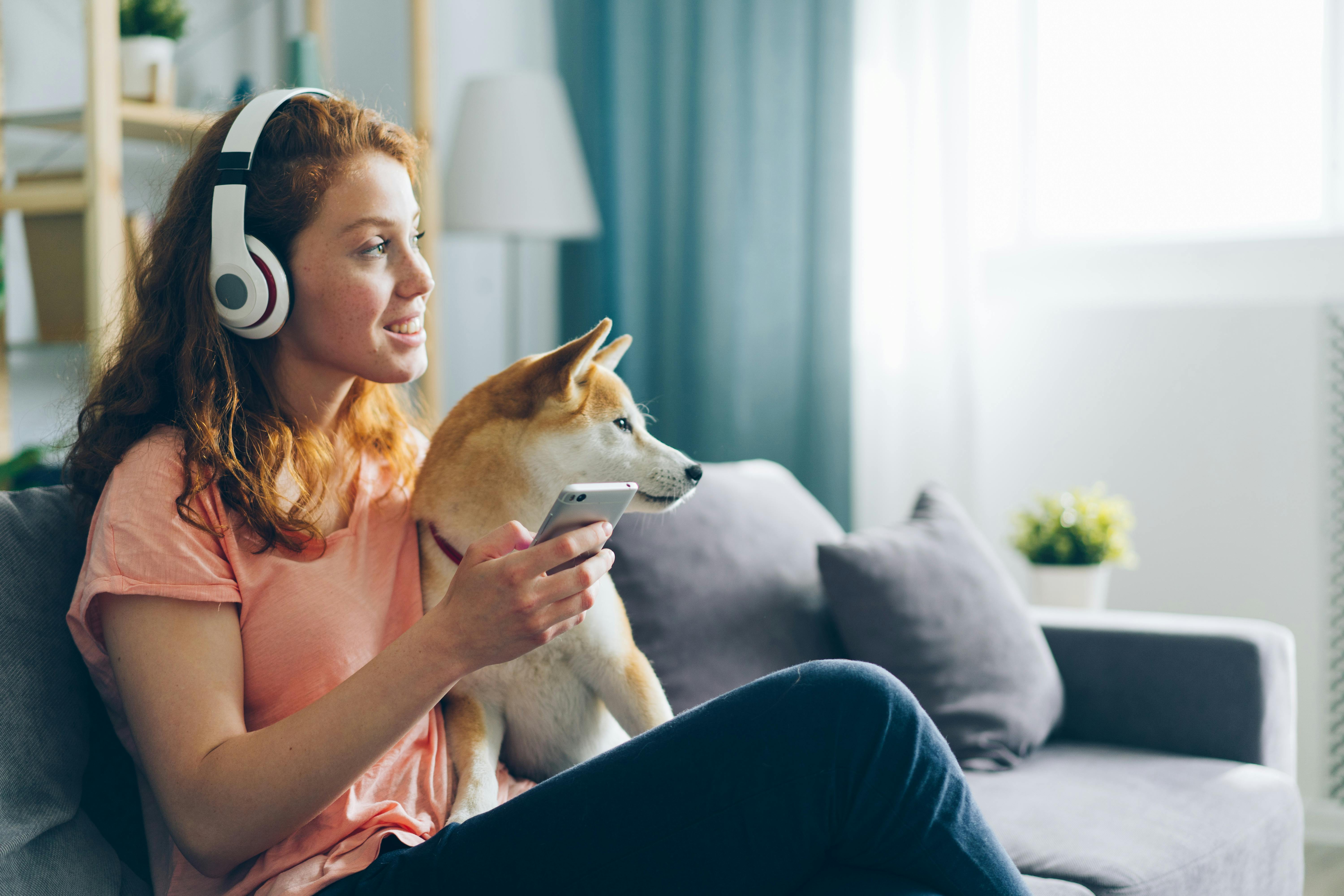 A Woman with Headphones Sitting on a Sofa with Her Shiba Inu Dog and Holding a Smartphone