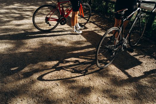 Two cyclists resting in a sun-dappled park in London, casting long shadows.