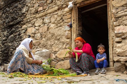 Free Two women and a child sit outside a rustic stone house, preparing vegetables. Stock Photo