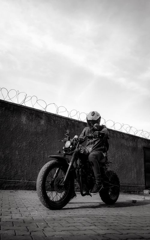 Free Black and white image of a motorcycle rider in full gear in front of a barbed wall on an urban street. Stock Photo