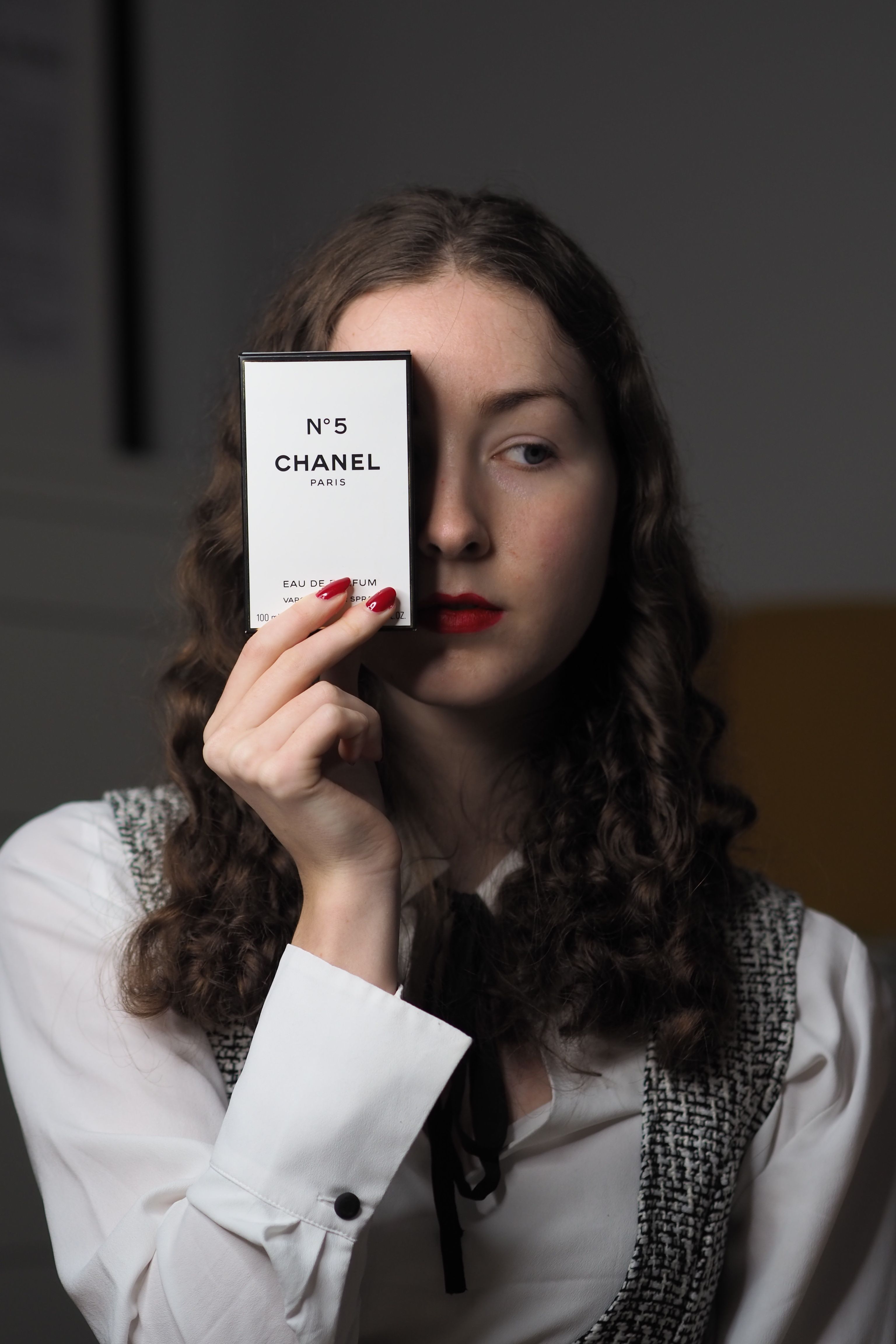 Portrait of a woman with curly hair holding Chanel No. 5 perfume elegantly indoors.