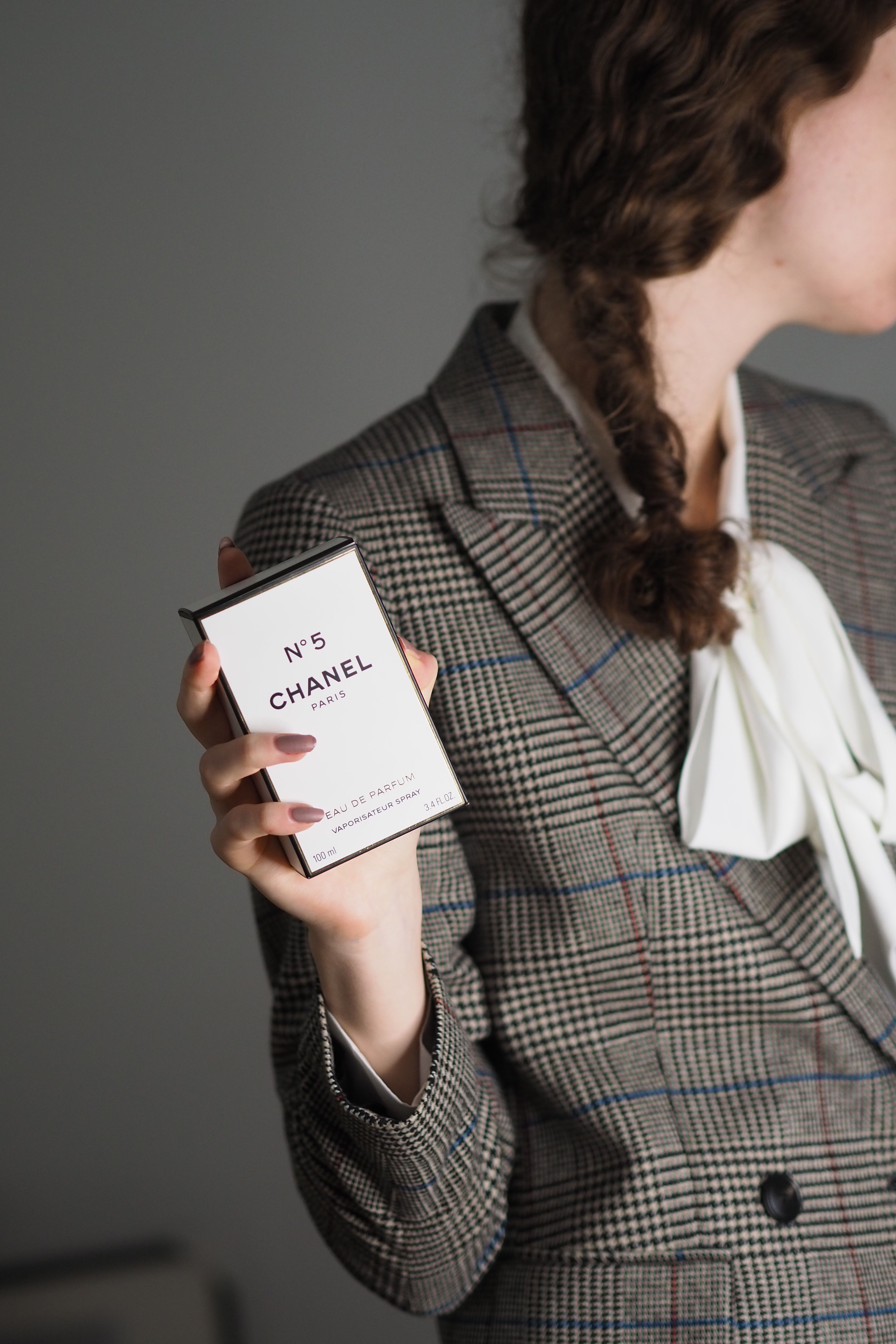Mulher estilosa com jaqueta xadrez segurando uma caixa de perfume Chanel No. 5 em ambiente interno.