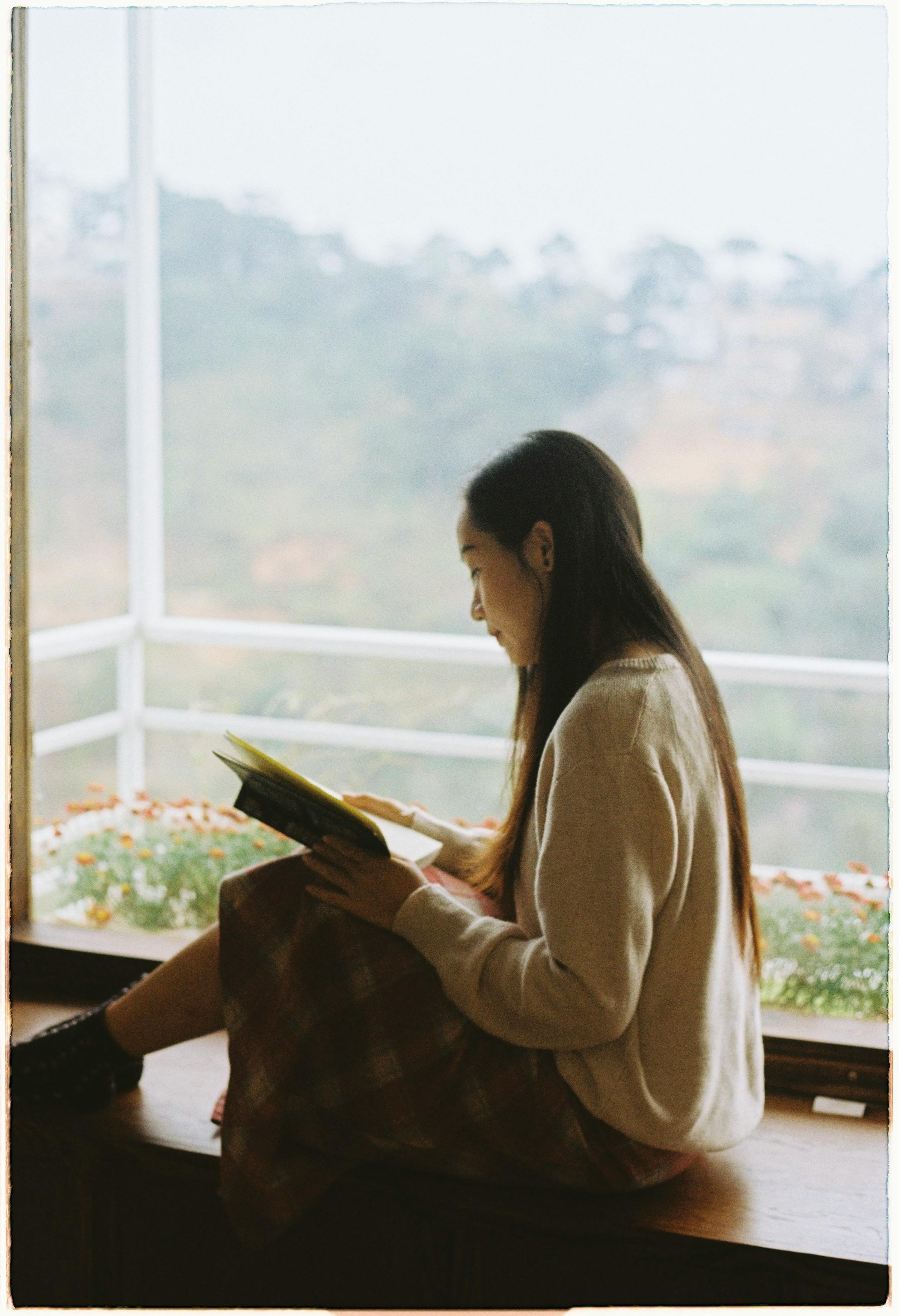 Young woman enjoying a book by the window with a serene mountain view.