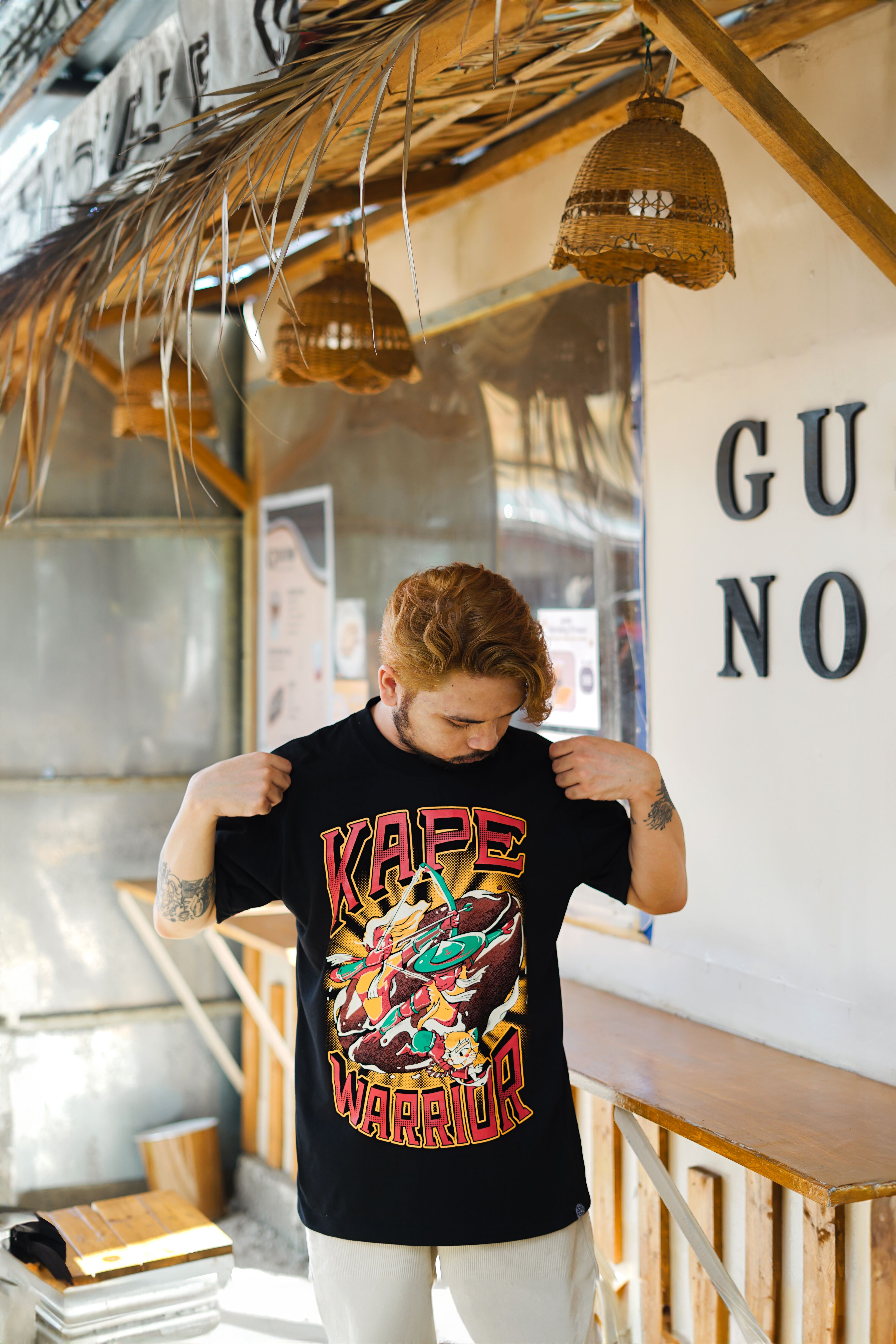 A young man wearing a graphic Kape Warrior t-shirt stands outdoors, checking his shirt design.