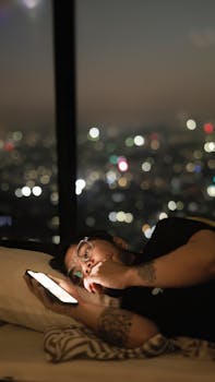 A man lying in bed using a smartphone at night, illuminated by city lights outside a window.
