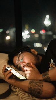 Man with eyeglasses using smartphone in bed at night with city lights.