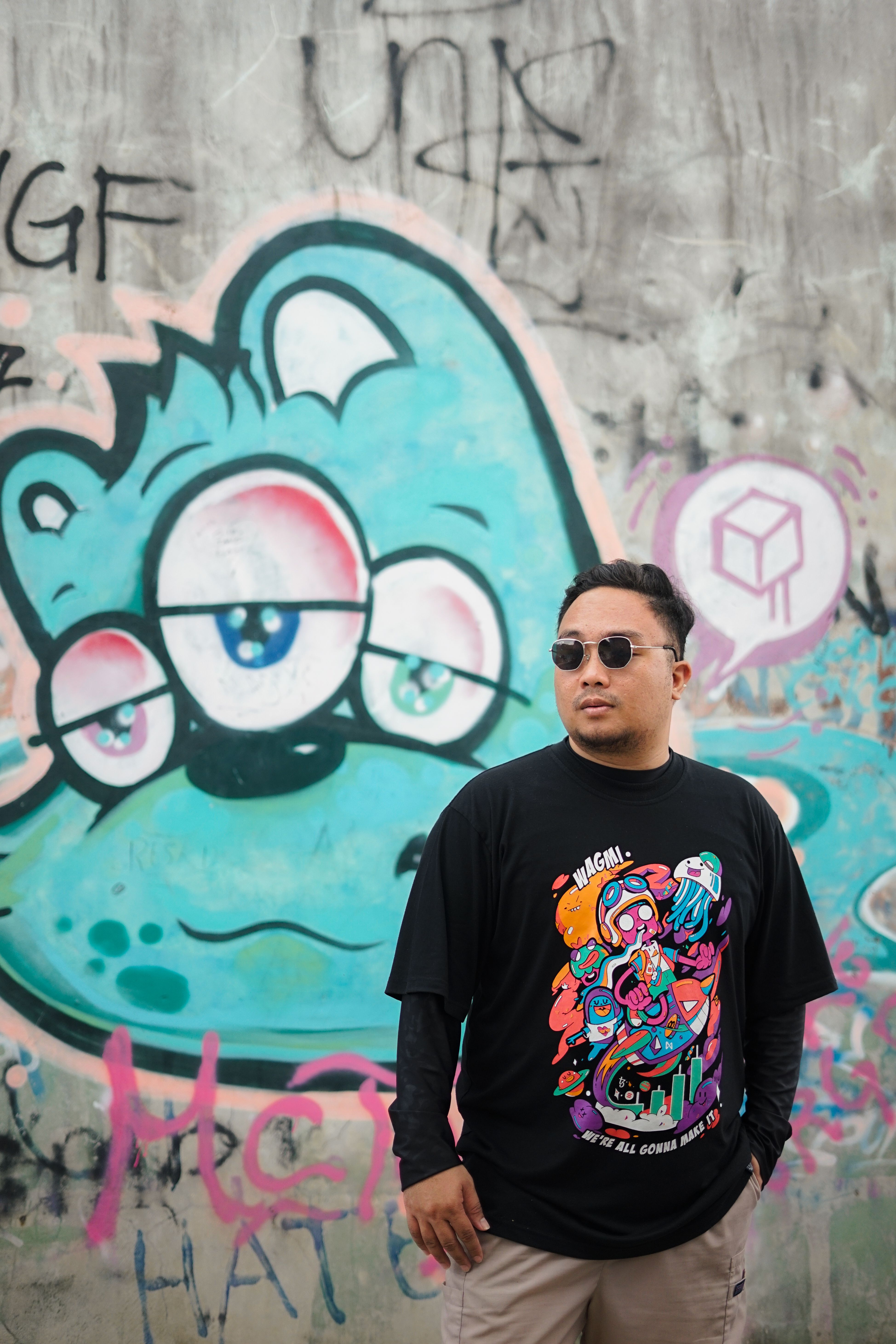 Asian man in sunglasses stands confidently against a vibrant graffiti wall in an urban setting.